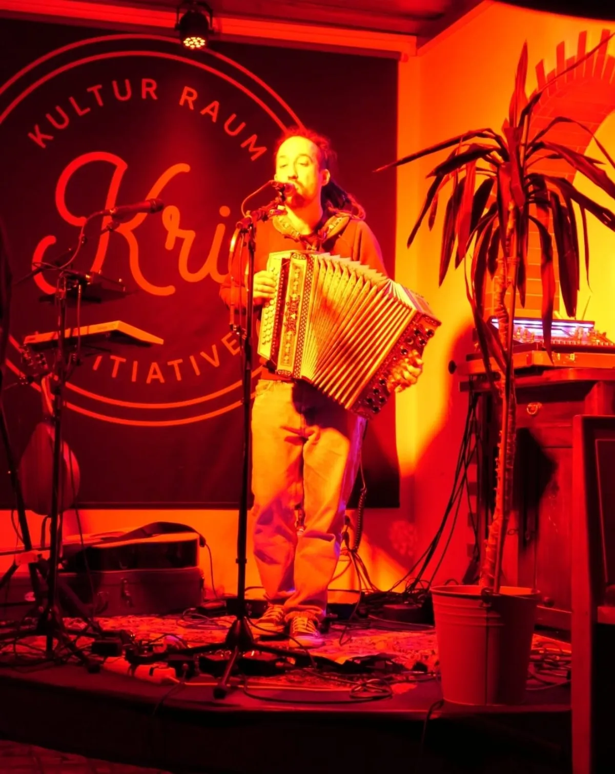 Beda solo auf der KRI-Bühne mit steirischer Harmonika, daneben eine Palme im orangeroten Licht