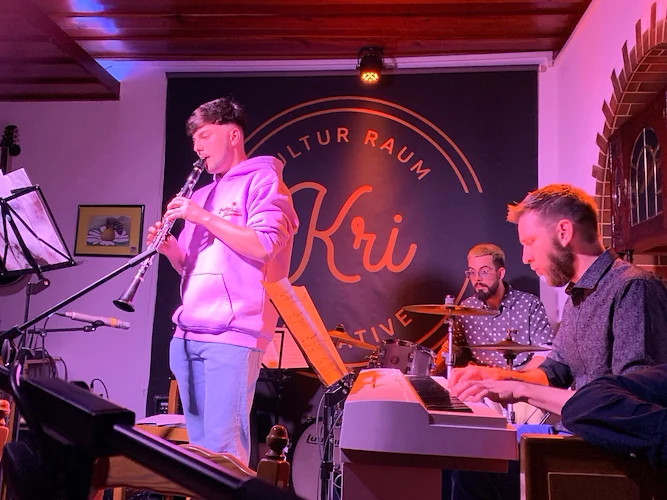 Klarinettist im rosa Hoodie auf der KRI-Bühne, dahinter Schlagzeuger und Keyboarder vor dem Kri-Banner